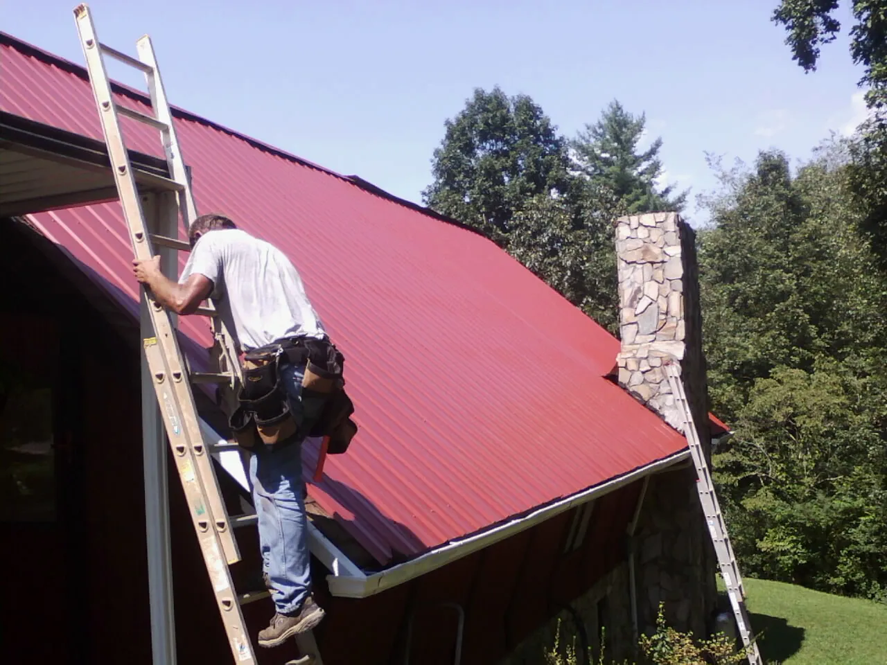 Roofer on a ladder beside a red metal roof and stone chimney — Soffit & Fascia Repair by Roofing Brothers in Bonita